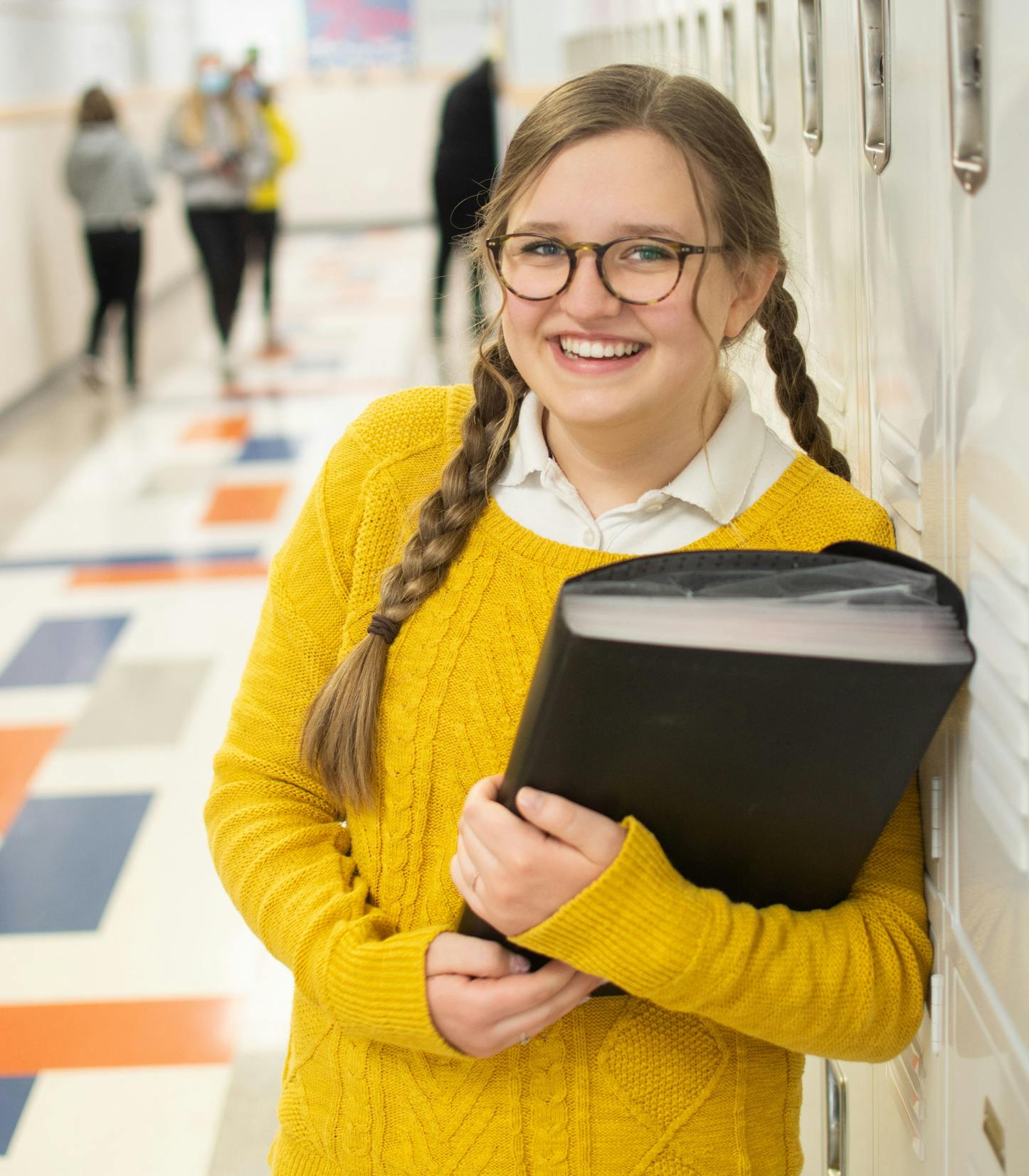 Female student smiling and standing against locker with work book.