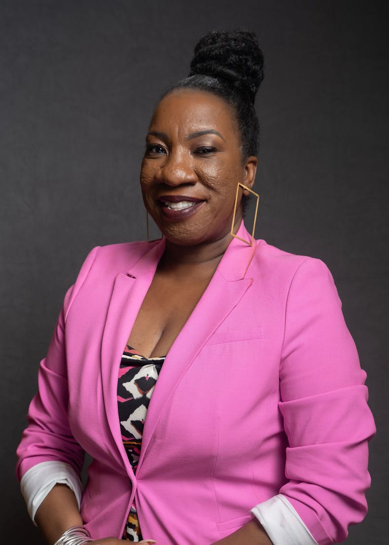  A photo of Tarana Burke, a Black woman, her hair is in a bun and she is wearing a pink blazer and patterned shirt, she has rectangular earrings and is smiling at the camera 